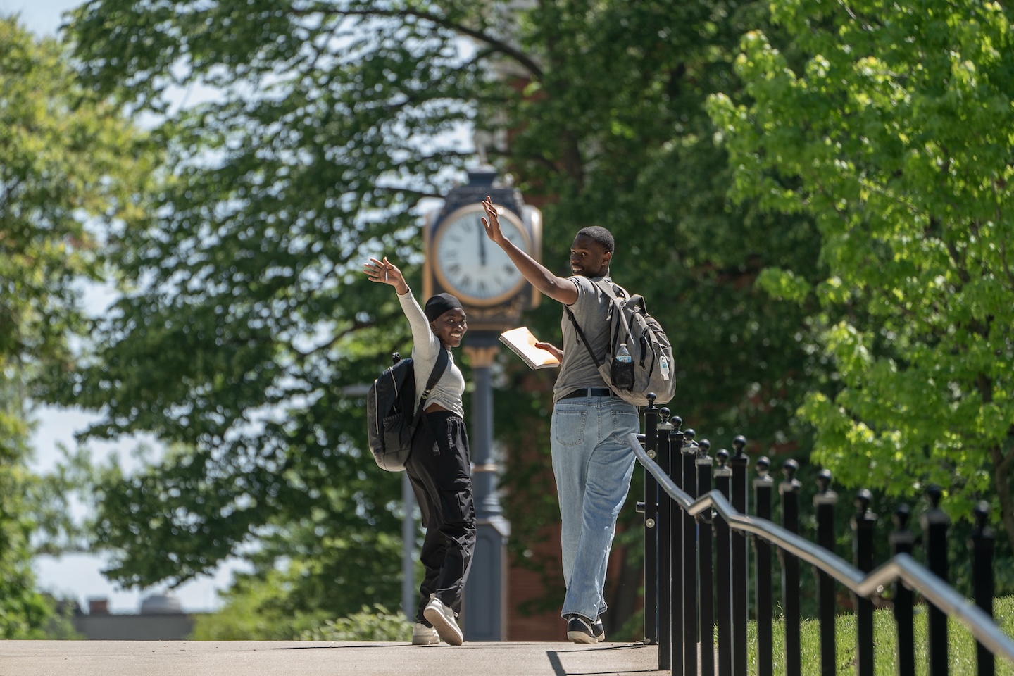 Two University of Akron students walk across campus on a sunny spring day, waving hello to someone off-camera, with backpacks and a textbook, surrounded by lush green trees and a campus clock tower in the background.